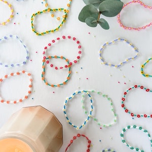 May include: A collection of colourful beaded bracelets with flower designs. The bracelets are arranged on a white surface with a candle in the bottom left corner.
