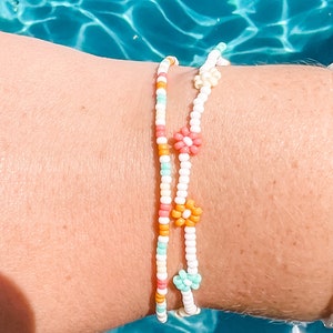 May include: Two beaded bracelets with colourful flower accents. The bracelets are white with orange, pink, and light blue beads. The flowers are made of pink, orange, and light blue beads.
