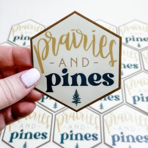 Pode incluir: Um adesivo hexagonal com contorno marrom e fundo branco. O adesivo apresenta o texto "prairies -AND- pines" em letras douradas e azul marinho. Um pequeno ícone de pinheiro fica abaixo do texto.