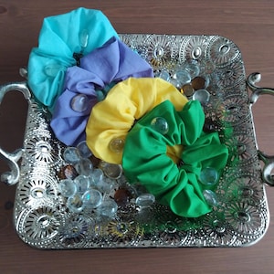 Op de afbeelding: Vier stoffen scrunchies in een zilveren dienblad met decoratieve uitsnijdingen. De scrunchies zijn turquoise, lavendel, geel en groen.