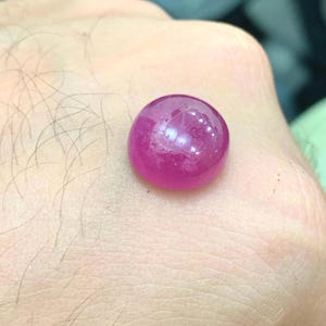 May include: A spherical, polished, deep pink gemstone. The gem has a smooth, reflective surface, with internal light reflections. The stone is held in a hand, showcasing its colour and shape. This could be a ruby or similar precious stone.