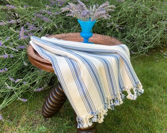 Puede incluir: Una manta crema con rayas verticales azules y ribete de borlas está sobre una pequeña mesa de madera oscura. Un jarrón turquesa contiene ramitas de lavanda. La mesa está sobre un césped verde, con arbustos de lavanda al fondo.