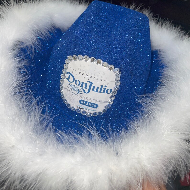 Puede incluir: Un sombrero vaquero azul con plumas blancas alrededor del borde. El sombrero tiene un logotipo de Don Julio Tequila con diamantes de imitaci&oacute;n.