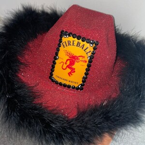 May include: Red glitter cowboy hat with a black feather trim. The hat features a yellow Fireball Cinnamon Whisky logo with a red devil and black rhinestones.
