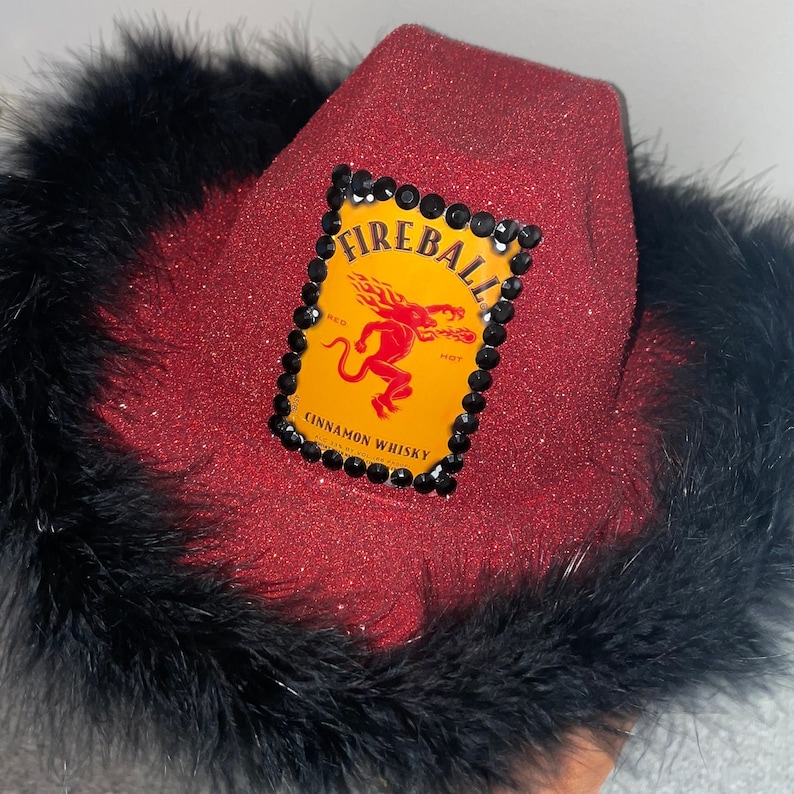 May include: Red glitter cowboy hat with a black feather trim. The hat features a yellow Fireball Cinnamon Whisky logo with a red devil and black rhinestones.