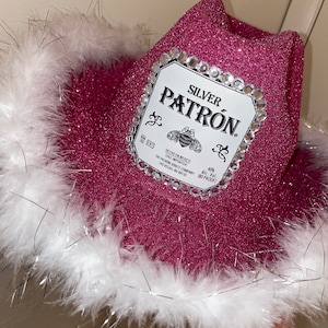 Puede incluir: Sombrero vaquero rosa con ribete de plumas blancas y una etiqueta de botella de tequila Patron plateada con pedrer&iacute;a. La etiqueta dice "SILVER PATRON. HECHO EN MEXICO SOLE IMPORTER THE PATRON SPIRITS COMPANY LAS VEGAS, NV 89118 40% Alc. Vol. (80 PROOF)"