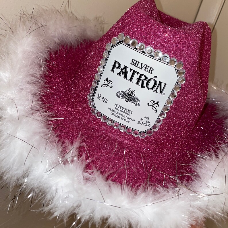 Puede incluir: Sombrero vaquero rosa con ribete de plumas blancas y una etiqueta de botella de tequila Patron plateada con pedrer&iacute;a. La etiqueta dice "SILVER PATRON. HECHO EN MEXICO SOLE IMPORTER THE PATRON SPIRITS COMPANY LAS VEGAS, NV 89118 40% Alc. Vol. (80 PROOF)"