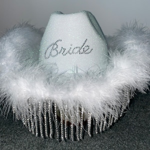 Puede incluir: Sombrero vaquero blanco con ribete de plumas blancas y flecos de pedrer&iacute;a plateada. El sombrero tiene la palabra "Bride" escrita en pedrer&iacute;a plateada en la parte delantera.