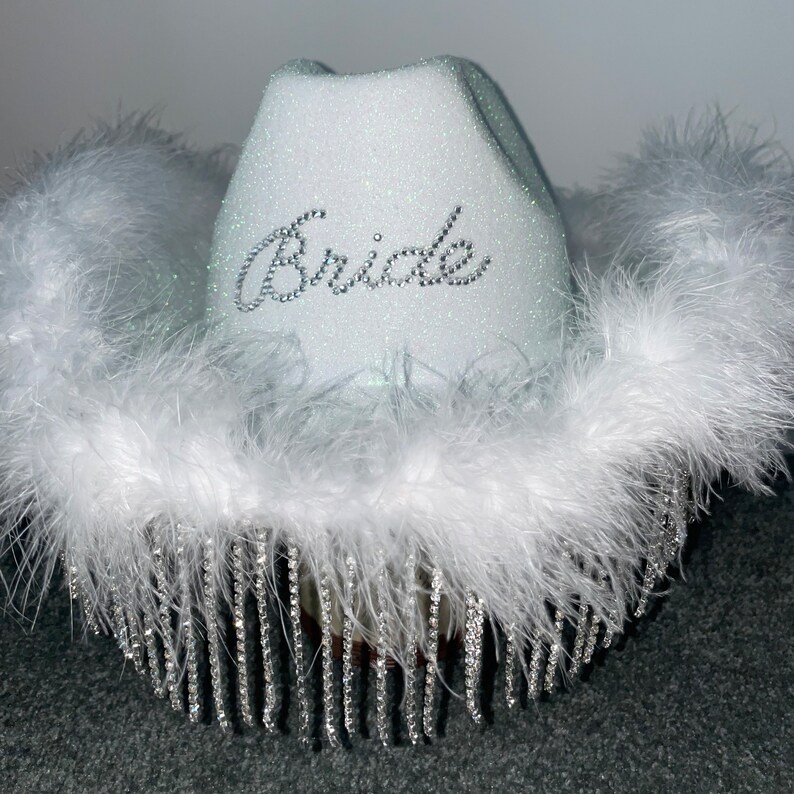 Puede incluir: Sombrero vaquero blanco con ribete de plumas blancas y flecos de pedrer&iacute;a plateada. El sombrero tiene la palabra "Bride" escrita en pedrer&iacute;a plateada en la parte delantera.