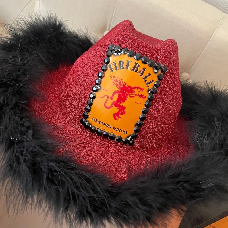 May include: A red cowboy hat with black feathers and a Fireball Cinnamon Whisky logo with rhinestones.