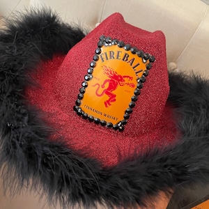 Puede incluir: Un sombrero vaquero de lentejuelas rojas con ribete de plumas negras. El sombrero presenta un logotipo de Fireball Cinnamon Whisky con un diablo rojo y las palabras "Fireball" y "Cinnamon Whisky".