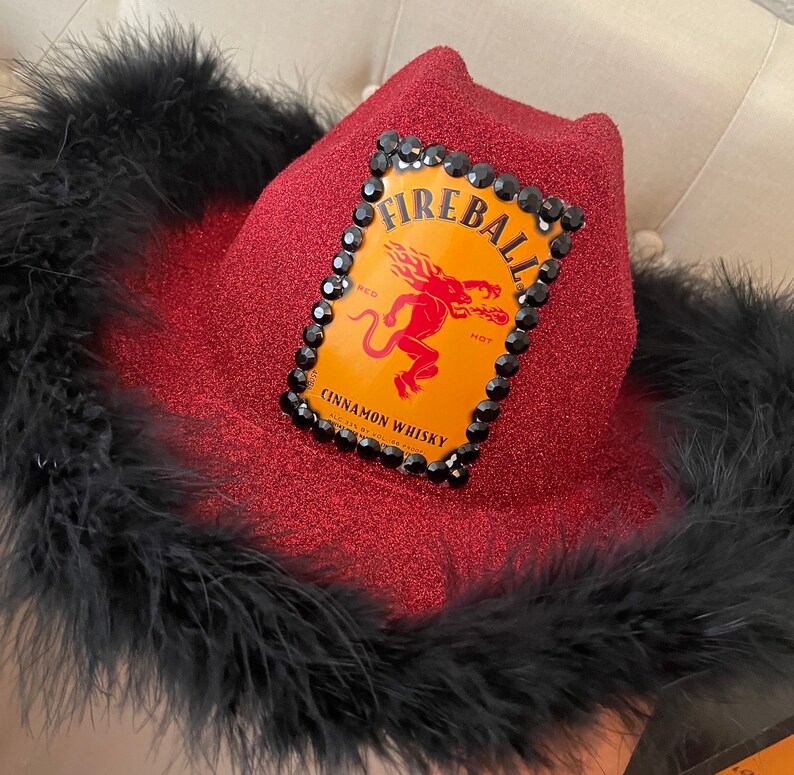 Puede incluir: Un sombrero vaquero de lentejuelas rojas con ribete de plumas negras. El sombrero presenta un logotipo de Fireball Cinnamon Whisky con un diablo rojo y las palabras "Fireball" y "Cinnamon Whisky".