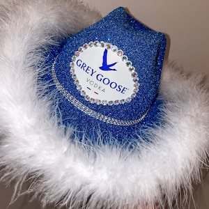 Puede incluir: Un sombrero vaquero azul con plumas blancas y un parche ovalado blanco con el texto "GREY GOOSE VODKA" y un logotipo de ganso volando.