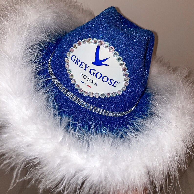 Puede incluir: Un sombrero vaquero azul con plumas blancas y un parche ovalado blanco con el texto "GREY GOOSE VODKA" y un logotipo de ganso volando.