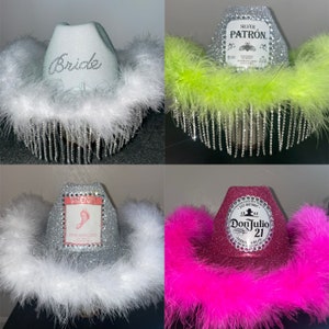 Puede incluir: Cuatro sombreros de vaquero plateados con diferentes adornos. Los sombreros est&aacute;n decorados con diamantes de imitaci&oacute;n y tienen plumas de varios colores. Los sombreros est&aacute;n etiquetados con las palabras "Bride", "Patron", "Pink Moscato" y "Don Julio 21".