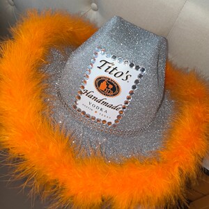 Puede incluir: Un sombrero vaquero de purpurina plateada con ribete de plumas naranja. El sombrero tiene una etiqueta de Tito's Handmade Vodka con diamantes de imitaci&oacute;n alrededor del borde.