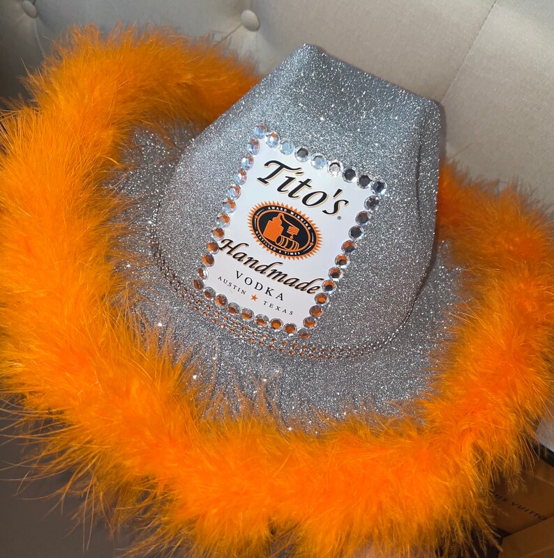 Puede incluir: Un sombrero vaquero de purpurina plateada con ribete de plumas naranja. El sombrero tiene una etiqueta de Tito's Handmade Vodka con diamantes de imitaci&oacute;n alrededor del borde.
