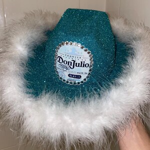 Puede incluir: Un sombrero vaquero de purpurina turquesa con ribete de plumas blancas. El sombrero tiene una etiqueta de Don Julio Tequila con pedrer&iacute;a.