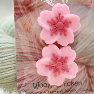 Könnte beinhalten: Zwei rosa, blütenförmige Seifen mit dunkleren rosa Zentren werden in einer transparenten Verpackung präsentiert. Die Verpackung befindet sich vor einem Wollknäuel. Der Text "Wooly Chicken" ist auf der Verpackung zu sehen.