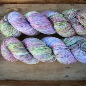 Könnte beinhalten: Zwei Stränge handgefärbter Wolle in einer Holzkiste. Das Garn ist eine Mischung aus Lavendel, Rosa und Grün, wodurch ein weiches, gesprenkeltes Aussehen entsteht. Ideal für Strick- oder Häkelprojekte.