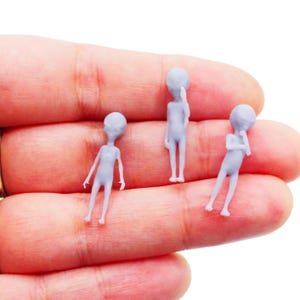 May include: Three small, light gray alien figurines. Each figurine has a large head and slender body. One figurine is standing, one is touching its head, and the third is leaning with a hand on its chin. The figurines are held in a hand.