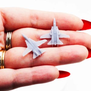 May include: Two small, light gray model fighter jets with wings and tails are displayed. The jets are resting on a surface. The jets are miniature replicas of a fighter jet.