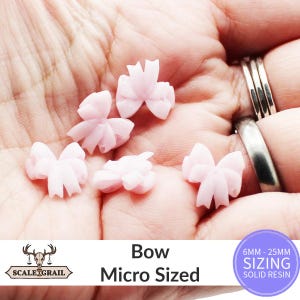 May include: Several micro-sized, light pink resin bows are displayed in a hand. The bows have a delicate, layered design. Text on the image reads "Bow Micro Sized" and "6MM - 25MM SIZING SOLID RESIN".