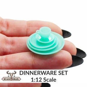 May include: A miniature teal dinnerware set, including a teacup, saucer, and plates. The set is 1:12 scale. The image shows a close-up of the set, highlighting the details of the tiny dishes. The text "DINNERWARE SET 1:12 Scale" is visible.
