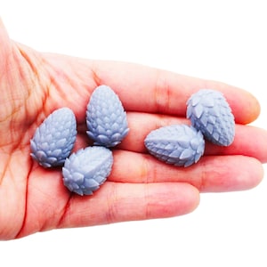 May include: Six small, light blue pine cone-shaped objects are held in a hand. The cones have a textured, scale-like surface. The objects are likely decorative or miniature items, possibly for crafts or home decor.