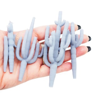 May include: Several light blue cactus figurines with detailed textures are held in a hand. The cacti vary in size and shape, with some having multiple arms. The background is plain white, highlighting the desert plant sculptures.
