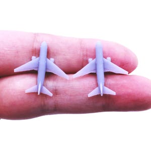 May include: A pair of small, light purple airplane-shaped stud earrings. Each earring features a detailed miniature aircraft design with wings, a fuselage, and a tail section, crafted in a solid, matte-finish material for a simple look.