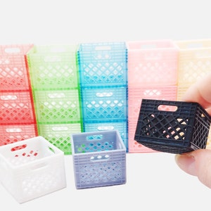 May include: A collection of miniature, colorful plastic crates in various colors, including red, green, blue, pink, and yellow. A hand holds a small black crate in the foreground. The crates have a lattice design and are stacked in rows.
