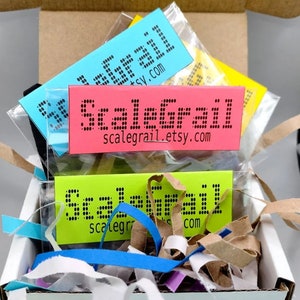 May include: Three small packages with the text "Scalegrail" and the website address "scalegrail.etsy.com" printed on them. The packages are filled with packing peanuts and are in a white box.