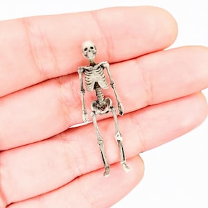 Może przedstawiać: Mała, szczegółowa miniaturowa figurka szkieletu o jasnoszarym, postarzanym wykończeniu kości z ciemnymi cieniowaniami w zagłębieniach, które podkreślają strukturę klatki piersiowej, czaszki, kręgosłupa oraz kończyn.