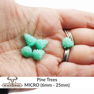 Puede incluir: Un puñado de pequeños pinos en miniatura de color verde claro. Los árboles en miniatura están agrupados y tienen una apariencia texturizada en forma de cono. La imagen incluye el texto "Pine Trees MICRO (6mm - 25mm)" y un logotipo.
