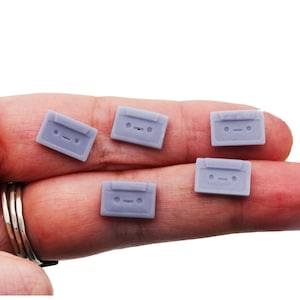 Puede incluir: Se muestran cinco pequeños amuletos de cinta de cassette de color púrpura claro. Cada amuleto tiene forma rectangular con un diseño detallado, que incluye una abertura central y dos agujeros circulares. Los amuletos se sostienen en una mano sobre un fondo blanco.
