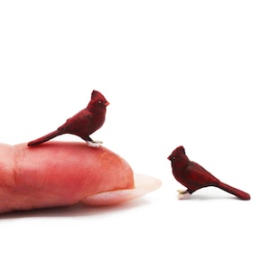 以下が含まれることがあります： 2つのミニチュアの赤いカージナルバードの置物。1羽の鳥は指先に止まり、もう1羽は近くに立っています。鳥は詳細な特徴があり、白い背景に対して足に白いアクセントがあります。