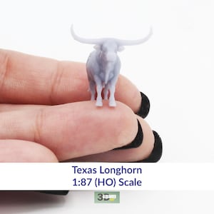 May include: A gray plastic model of a Texas Longhorn bull in 1:87 (HO) scale. The model is standing on a hand.