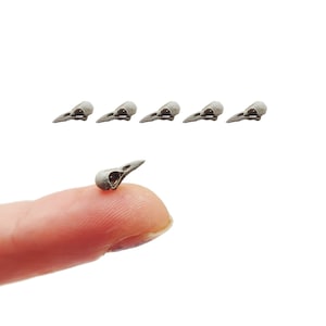 May include: A collection of six miniature silver-toned bird skull charms. Each charm features a detailed, hollowed-out eye socket and a pointed beak. The small, metallic pieces are arranged in a row above a single skull resting on a fingertip.