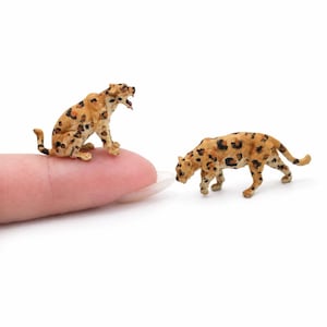 Pode incluir: Duas figuras em miniatura de leopardos com pelagem castanha e manchas pretas. Um leopardo está sentado com a boca aberta, enquanto o outro está de pé numa postura de espreita. Ambas as figuras são pequenas o suficiente para caber num dedo.