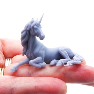 May include: A small, light gray unicorn figurine in a resting position. The creature features a spiraled horn on its forehead, a flowing mane, and a long tail. The surface of the figurine is smooth and matte, showing detailed muscle definition.