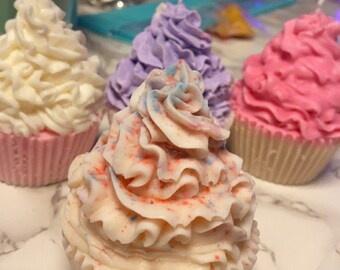 May include: A collection of cupcake-shaped candles with white, pink, and purple frosting. The candles have a realistic design, with detailed frosting swirls and colorful sprinkles. The candles are on a marble surface.