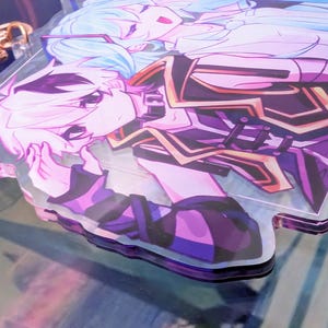 Pode incluir: Um porta-chaves de acr&iacute;lico transparente com uma ilustra&ccedil;&atilde;o impressa de duas personagens de estilo anime com cabelo azul claro e branco. As personagens vestem trajes roxo-escuro e preto com detalhes dourados. Um fecho de metal em forma de estrela em ouro rosa est&aacute; fixado.