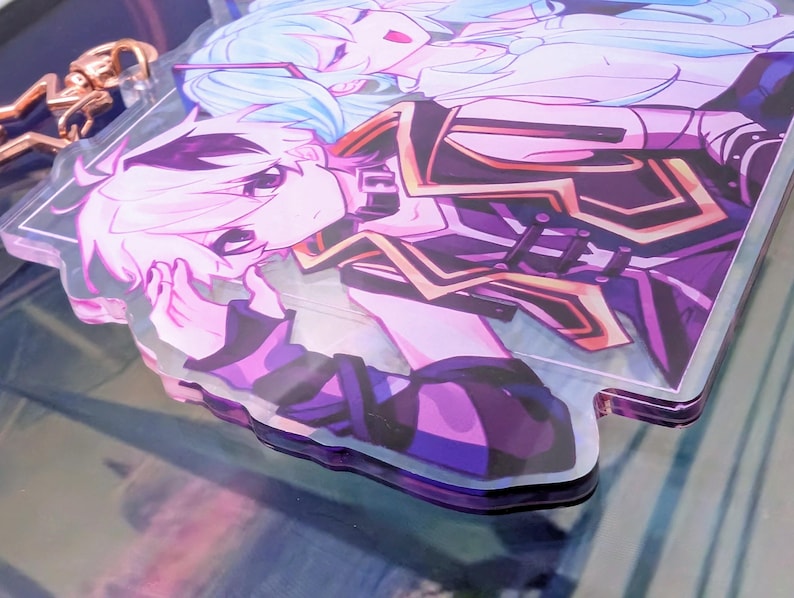 Pode incluir: Um porta-chaves de acr&iacute;lico transparente com uma ilustra&ccedil;&atilde;o impressa de duas personagens de estilo anime com cabelo azul claro e branco. As personagens vestem trajes roxo-escuro e preto com detalhes dourados. Um fecho de metal em forma de estrela em ouro rosa est&aacute; fixado.