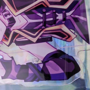 Pode incluir: Um expositor de acr&iacute;lico transparente com uma personagem de estilo anime de cabelo rosa claro, vestindo um traje roxo escuro e preto com detalhes dourados. A personagem &eacute; representada do peito para cima, com uma m&atilde;o perto do queixo.