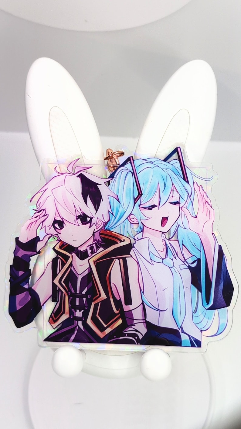 Pode incluir: Um pingente de acr&iacute;lico transparente com dois personagens em estilo anime. Um personagem tem cabelo branco curto com detalhes pretos e veste um colete preto. O outro personagem tem cabelo longo azul claro e veste uma camisa branca com gravata azul.