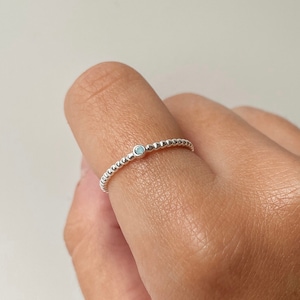 May include: A dainty silver ring featuring a small, pale blue gemstone set in the centre. The ring is crafted from tiny beads, giving it a textured appearance.