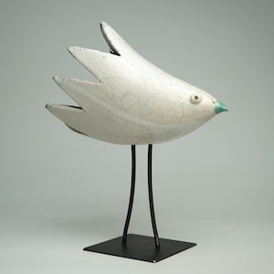 May include: A ceramic bird sculpture with a white crackle glaze finish and a small turquoise beak. The bird has stylized, pointed wing feathers and is mounted on two thin, black metal legs attached to a square black metal base.