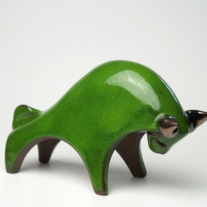 Op de afbeelding: Een keramieken stierfiguur in een levendige groene glazuur. De sculptuur heeft een gestileerd ontwerp met een gebogen lichaam, bruine poten en donkerbruine hoorns. De stier is een decoratief item.