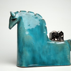 Può includere: Scultura di cavallo in ceramica turchese con finitura lucida. Il cavallo ha una forma stilizzata con un lungo collo e una piccola coda. Due piccole case scure sono sulla sua schiena.
